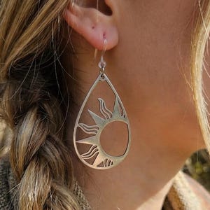 May include: Silver-toned teardrop earrings featuring a sun design. The earrings have a cut-out sun with rays and a circular center. The earrings are lightweight and dangle from a simple hook.