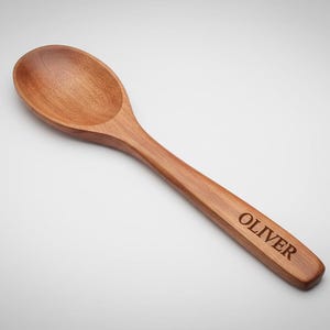 K&ouml;nnte beinhalten: Ein Holzl&ouml;ffel mit einem abgerundeten L&ouml;ffel und einem langen Griff. Der Griff ist mit dem Namen "OLIVER" graviert. Der L&ouml;ffel ist aus hellbraunem Holz gefertigt und wird wahrscheinlich zum Kochen oder Servieren verwendet.