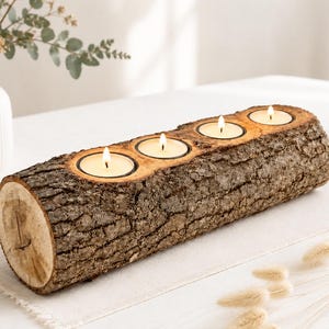 Könnte beinhalten: Ein zylindrischer Kerzenhalter aus einem Holzstamm mit rauer, strukturierter brauner Rinde an der Außenseite. Die Oberseite enthält vier kreisförmige Aussparungen, in denen jeweils ein kleines, brennendes weißes Teelicht steht.