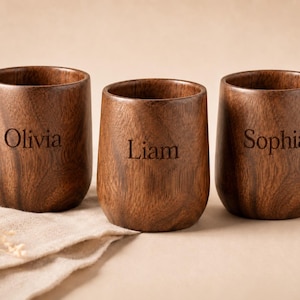Può includere: Tre bicchieri di legno con nomi incisi: Olivia, Liam e Sophia. I bicchieri di legno scuro hanno una finitura liscia e sono esposti su un panno beige. Lo sfondo è di colore neutro.
