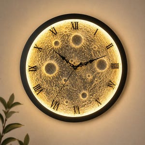 Puede incluir: Un reloj de pared redondo con un marco negro y una esfera luminosa con forma de luna. El reloj presenta números romanos y manecillas negras. La esfera lunar tiene cráteres y un brillo amarillo cálido, creando una pieza decorativa única.