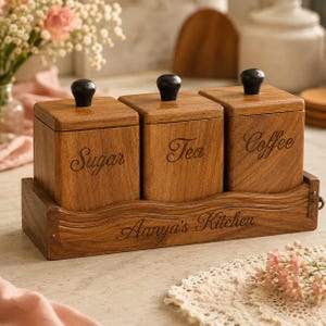 Könnte beinhalten: Ein Küchenbehälter-Set aus Holz mit drei quadratischen Behältern mit den Aufschriften "Sugar", "Tea" und "Coffee". Jeder Behälter hat einen schwarzen Knopf oben und steht auf einem Holztablett mit der Gravur "Aanya's Kitchen".