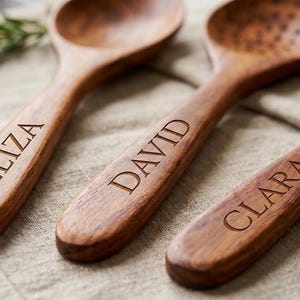 K&ouml;nnte beinhalten: Drei h&ouml;lzerne K&uuml;chenutensilien: ein L&ouml;ffel, ein Schlitzl&ouml;ffel und eine Gabel. Jedes Utensil ist mit einem Namen graviert: Eliza, David und Clara. Die Utensilien sind aus warmbraunem Holz und liegen auf einem beigefarbenen Leinentuch.