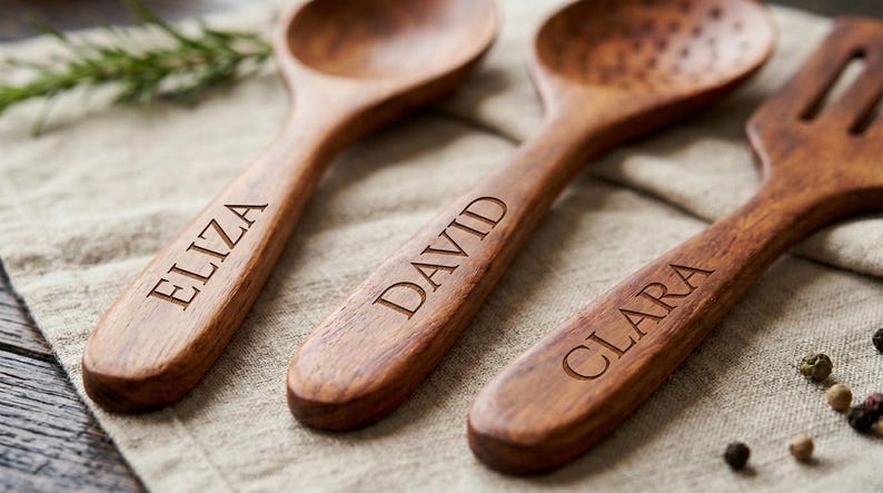 K&ouml;nnte beinhalten: Drei h&ouml;lzerne K&uuml;chenutensilien: ein L&ouml;ffel, ein Schlitzl&ouml;ffel und eine Gabel. Jedes Utensil ist mit einem Namen graviert: Eliza, David und Clara. Die Utensilien sind aus warmbraunem Holz und liegen auf einem beigefarbenen Leinentuch.