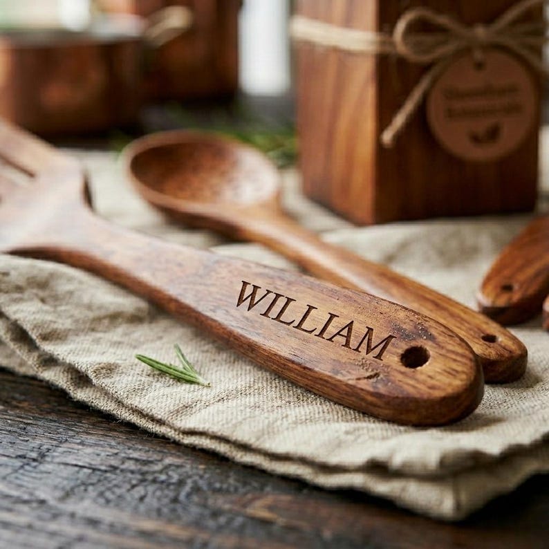 K&ouml;nnte beinhalten: K&uuml;chenutensilien aus Holz, darunter eine Gabel und ein L&ouml;ffel, sind mit dem Namen "WILLIAM" graviert. Die Utensilien sind aus dunklem Holz gefertigt und liegen auf einem beigen Leinentuch. Im Hintergrund befindet sich eine Holzkiste.