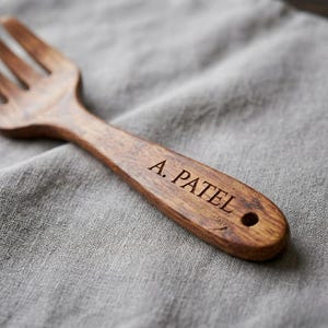 K&ouml;nnte beinhalten: Eine Holzgabel mit dunkelbraunem Finish, graviert mit dem Namen "A. Patel". Die Gabel liegt vor einem grauen Leinenhintergrund. Der Griff der Gabel hat ein Loch am Ende.