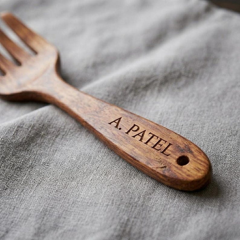 K&ouml;nnte beinhalten: Eine Holzgabel mit dunkelbraunem Finish, graviert mit dem Namen "A. Patel". Die Gabel liegt vor einem grauen Leinenhintergrund. Der Griff der Gabel hat ein Loch am Ende.