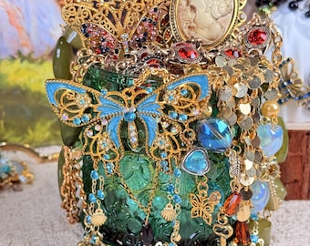 Peut inclure: Un assortiment de bijoux ornés, comprenant des colliers, des boucles d'oreilles et des broches, débordant d'un bocal en verre vert. Les bijoux présentent des filigranes dorés, des pierres précieuses colorées et des motifs de papillons. Une camée et d'autres éléments décoratifs sont également visibles.