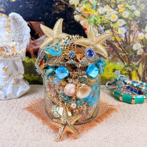 Puede incluir: Un tarro de cristal lleno de varias joyas y objetos decorativos. El tarro está adornado con estrellas de mar doradas, gemas azules y conchas marinas. El contenido incluye anillos, collares y otras baratijas, creando un tema costero o náutico.