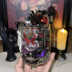 Puede incluir: Un tarro transparente lleno de diversas joyas y accesorios de temática gótica. El tarro contiene collares, pendientes y broches en tonos rojos, negros y plateados. Un broche de mariposa roja y un dije morado con forma de ojo son visibles. El tarro se sostiene en una mano.