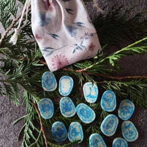Peut inclure: Ensemble de pierres runiques bleues et blanches avec divers symboles, disposées sur un lit de feuillage vert. Un sac à cordon floral est posé au-dessus des pierres. Le sac présente des motifs de fleurs roses et bleues.