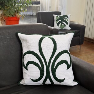 Puede incluir: Cojín cuadrado blanco con un diseño floral verde oscuro. El cojín tiene una textura acolchada y un borde verde oscuro. Otro cojín con un diseño de hoja verde es visible en el fondo. Los cojines están sobre un sofá gris oscuro.