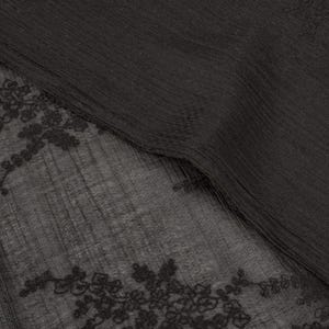 May include: A close-up shot of a black garment with a ribbed texture. The lower section has a sheer panel with detailed floral lace. The fabric looks soft and drapes, creating a contrast between the opaque and transparent areas.