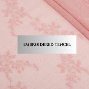 May include: Close-up of pink embroidered tencel fabric. The fabric features a delicate floral pattern embroidered in a matching pink thread. The text "EMBROIDERED TENCEL" is displayed on a rectangular label in the centre of the image.