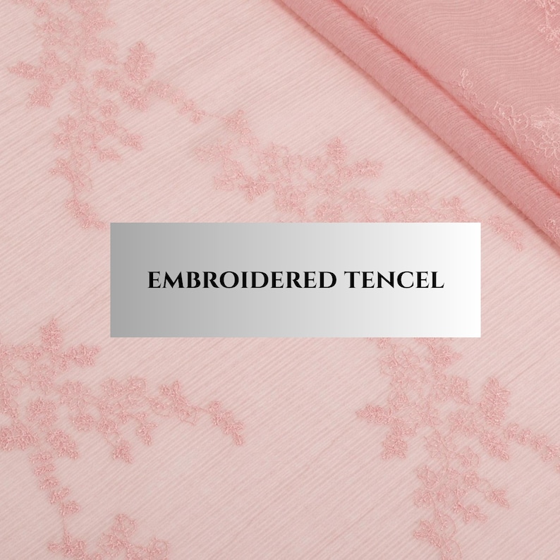 May include: Close-up of pink embroidered tencel fabric. The fabric features a delicate floral pattern embroidered in a matching pink thread. The text "EMBROIDERED TENCEL" is displayed on a rectangular label in the centre of the image.