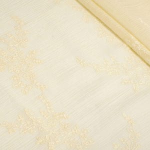 May include: Cream-coloured fabric with a delicate, textured surface. The fabric features intricate, embroidered floral patterns in a lighter shade of cream. The material is folded, revealing the layers and texture of the fabric.