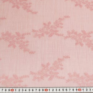 May include: Pale pink lace fabric with embroidered floral patterns. The fabric has a textured appearance and a ruler is visible along the bottom edge, showing measurements in centimetres.