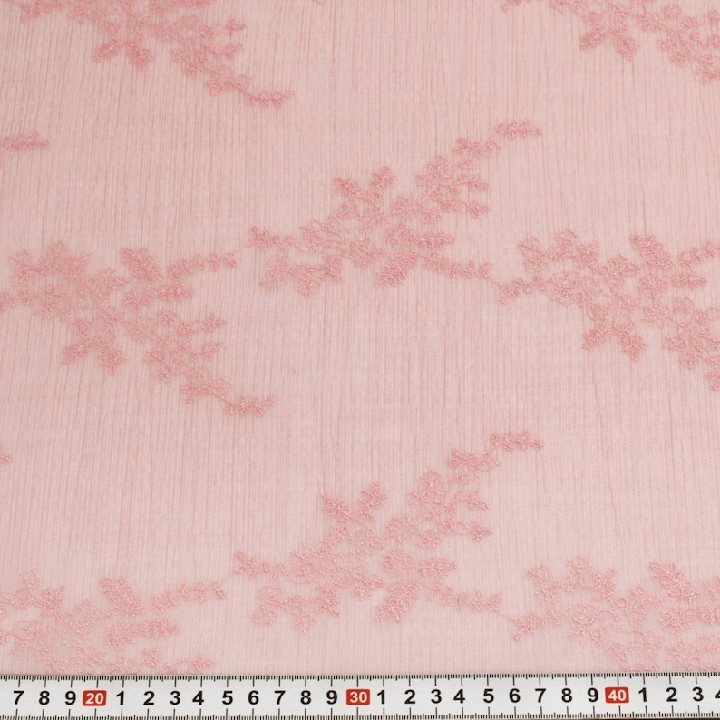 May include: Pale pink lace fabric with embroidered floral patterns. The fabric has a textured appearance and a ruler is visible along the bottom edge, showing measurements in centimetres.