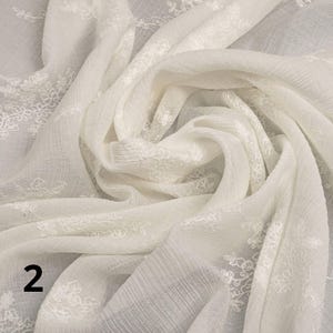 May include: Cream-coloured fabric with intricate floral embroidery. The fabric is draped and folded, creating soft shadows and textures. The embroidery is concentrated along the edges, adding a touch of elegance to the textile. The number "2" is visible in the lower left corner.