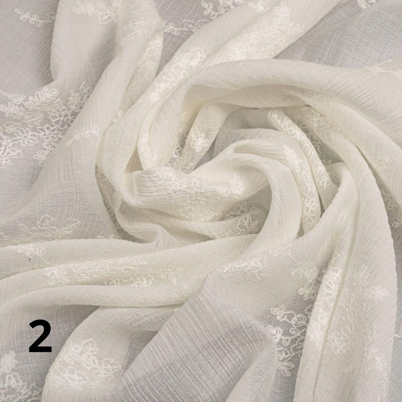 May include: Cream-coloured fabric with intricate floral embroidery. The fabric is draped and folded, creating soft shadows and textures. The embroidery is concentrated along the edges, adding a touch of elegance to the textile. The number "2" is visible in the lower left corner.