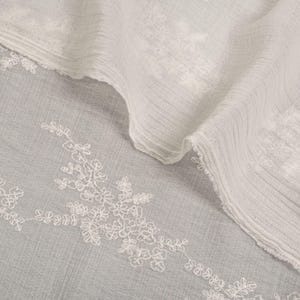 May include: Close-up of white fabric with floral embroidery. The fabric has a textured appearance with subtle lines and folds. The embroidery adds a touch of elegance. The overall aesthetic is soft and airy, suitable for clothing or home decor.