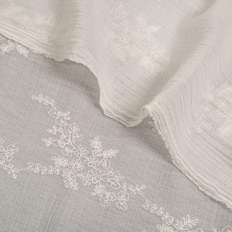 May include: Close-up of white fabric with floral embroidery. The fabric has a textured appearance with subtle lines and folds. The embroidery adds a touch of elegance. The overall aesthetic is soft and airy, suitable for clothing or home decor.