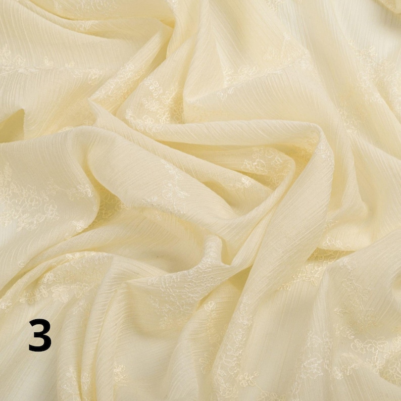 May include: A close-up shot of a cream-coloured fabric with a delicate floral pattern. The fabric is draped, creating soft folds and shadows. The number "3" is visible in the lower left corner. The fabric has a subtle sheen, suggesting a lightweight material.