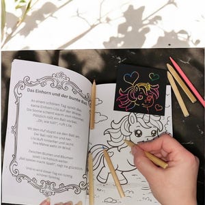 Op de afbeelding: Een kleurboek met een eenhoornthema, open op een pagina met een lijntekening van een eenhoorn. Er liggen verschillende kleurpotloden rond het boek. Een hand houdt een potlood vast en kleurt de eenhoorn in.