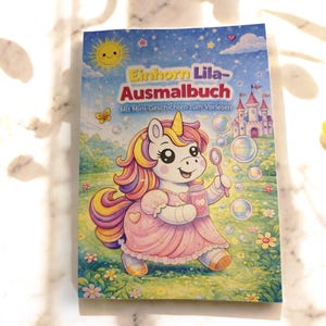 Op de afbeelding: Een kleurboek voor kinderen getiteld "Einhorn Lila-Ausmalbuch" met een cartoon eenhoorn die bellen blaast. De cover bevat een roze jurk, een kasteel en een zonnige hemel met sterren en een vlinder. Het boek ligt op een wit marmeren oppervlak.
