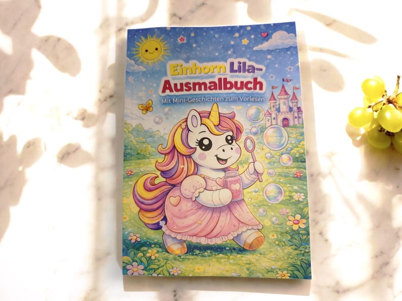 Op de afbeelding: Een kleurboek voor kinderen getiteld "Einhorn Lila-Ausmalbuch" met een cartoon eenhoorn die bellen blaast. De cover bevat een roze jurk, een kasteel en een zonnige hemel met sterren en een vlinder. Het boek ligt op een wit marmeren oppervlak.