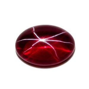 May include: A polished, oval-shaped, deep red gemstone with a star effect. The gem has a smooth surface and a bright, six-rayed star pattern radiating from the center. The stone is set against a white background.