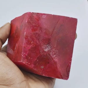 May include: A large, roughly-cut, ruby-red gemstone cube. The stone has a translucent quality, with visible internal textures and variations in color. The cube is held in a hand, showcasing its size and shape. The overall appearance suggests a raw, natural mineral specimen.