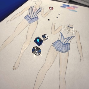 May include: A fashion sketch on paper showing two leotard designs in blue and white with intricate detailing, accompanied by three loose faceted crystals in blue and clear tones placed on the page near the illustrated figures.