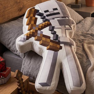 May include: A Minecraft-themed pillow in the shape of a skeleton character, holding a pixelated bow and arrow. The pillow is primarily white and gray, with brown and tan accents. It is resting on a gray bedspread.