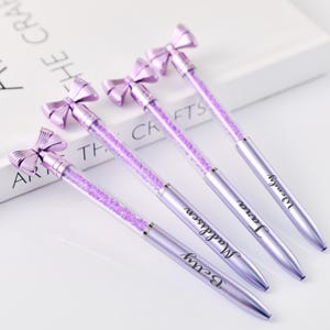 Może przedstawiać: Pięć lawendowych długopisów z wypełnionymi kryształami korpusami i kokardkami. Każdy długopis ma srebrną podstawę i jest spersonalizowany imieniem. Długopisy ułożone są na białej powierzchni.