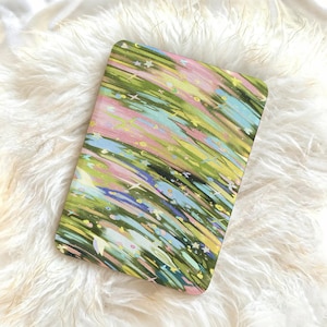 Puede incluir: Una funda rectangular para tableta con un patrón abstracto de pinceladas diagonales en tonos verde oliva, azul claro y rosa suave. El diseño está acentuado con pequeñas formas blancas de estrellas y corazones dispersas.