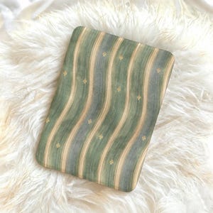 Op de afbeelding: Een rechthoekig decoratief kussen met een gestreept patroon in groen, beige en grijs. Kleine gouden bloemmotieven zijn gelijkmatig verdeeld over de strepen. Het kussen ligt op een pluizige witte achtergrond.