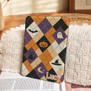 May include: A rectangular e-reader case featuring an argyle pattern in black, orange, and purple. The design includes small icons of a bat, ghost, pumpkin, black cat, witch hat, and crescent moon. The name Lucy is printed in white with an orange outline.