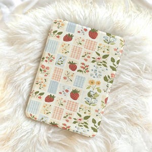 Könnte beinhalten: Ein rechteckiges, cremefarbenes Etui mit einem Patchwork-Design aus Erdbeeren, Blumen und karierten Quadraten in Rot, Rosa, Blau und Gelb. Das Etui ist vor einem flauschigen, weißen Hintergrund platziert.