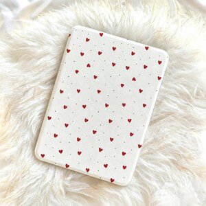May include: A rectangular, off-white phone case featuring a pattern of small red hearts and tiny black dots. The case has rounded corners and a smooth surface, creating a simple and charming design.
