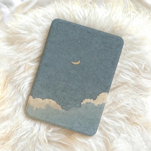 May include: A rectangular e-reader case with a matte blue-grey finish featuring a speckled texture. The design displays a small, light-colored crescent moon centered in the upper portion and a row of fluffy, light-colored clouds along the bottom edge.