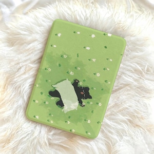 Puede incluir: Funda de teléfono rectangular con fondo verde claro que presenta un gato negro tumbado boca arriba con una almohada blanca. La funda tiene un estampado floral con pequeñas flores blancas y hojas verdes.