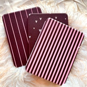 May include: Three rectangular cases with different patterns. One has vertical pink stripes on a dark background, another has vertical pink stripes, and the third has white hearts on a dark background.