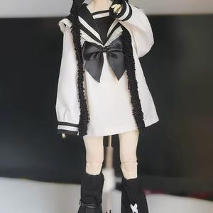 May include: A doll dressed in a white sailor-style outfit with black accents, including a large bow, trim, and boots. The doll wears a black bear-eared hat with a white cross. The doll's hair is white.