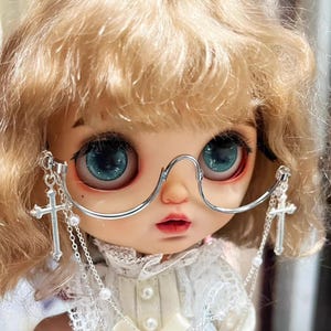Puede incluir: Una muñeca con grandes ojos azules y cabello rubio lleva gafas con montura plateada con dijes de cruz colgantes. La muñeca está vestida con un atuendo de encaje blanco, con un collar de perlas. Los labios de la muñeca son de un rojo suave.