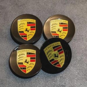 May include: Four black Porsche wheel center caps. Each cap features the Porsche crest: a yellow shield with a black horse, red and black stripes, and the word "PORSCHE" in gold lettering.
