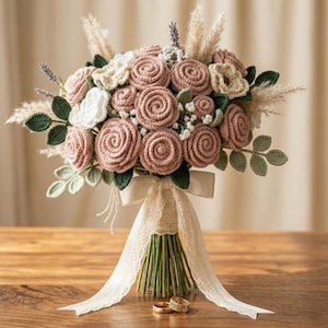 May include: A handcrafted crochet bouquet featuring dusty rose-colored roses, white and cream-colored flowers, and green leaves. The bouquet is tied with a cream-colored ribbon and sits on a wooden surface. Two gold wedding rings are at the base.