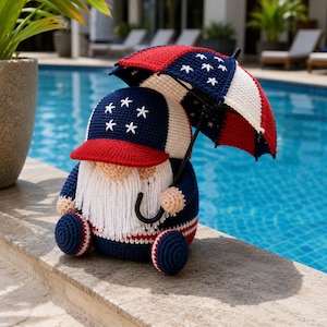 May include: A crocheted gnome figurine wearing a blue, white, and red baseball cap with white star embroidery. The gnome has a long white yarn beard and holds a small, matching red, white, and blue crocheted umbrella over its head.