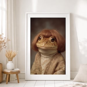 Puede incluir: Impresión artística enmarcada que presenta una rana con peluca y suéter. La rana tiene una tez marrón claro y lleva una peluca de color marrón rojizo. La rana lleva un suéter beige y un cuello alto blanco. El arte se muestra en un marco blanco.