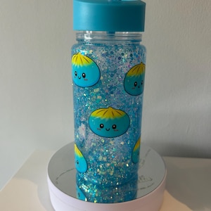 May include: A clear plastic tumbler with a blue lid and straw, filled with blue liquid and iridescent glitter. The exterior features a repeating pattern of small, light blue cartoon dumplings with yellow tops and smiling faces printed on the surface.
